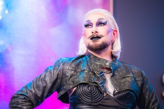 Drag Queen mit rosa Haaren und schwarzer Lederjacke vor buntem Hintergrund