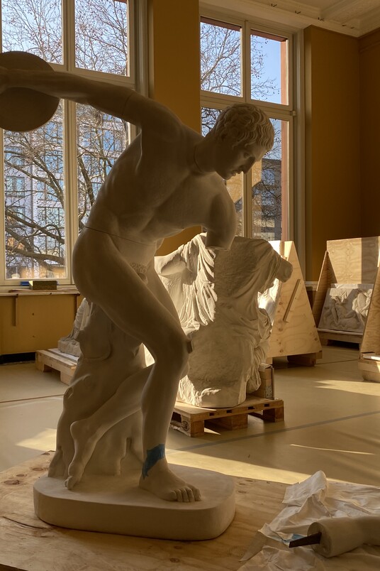 Im Vordergrund steht die Gipsfigur eines Diskuswerfers in einem hellen Raum, in dem auch andere Gipsfiguren gerade aus Boxen ausgepackt werden.