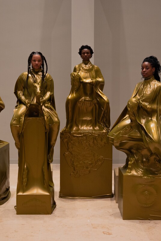 Sechs Frauen in goldenen Kleidern posieren auf großen würfelförmigen Objekten in einer Kunstgalerie