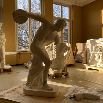 Im Vordergrund steht die Gipsfigur eines Diskuswerfers in einem hellen Raum, in dem auch andere Gipsfiguren gerade aus Boxen ausgepackt werden.