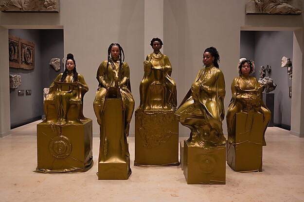 Sechs Frauen in goldenen Kleidern posieren auf großen würfelförmigen Objekten in einer Kunstgalerie